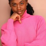 Woman in a bright pink turtleneck sweater smiles softly with her hand resting under her chin; neutral peach backdrop.