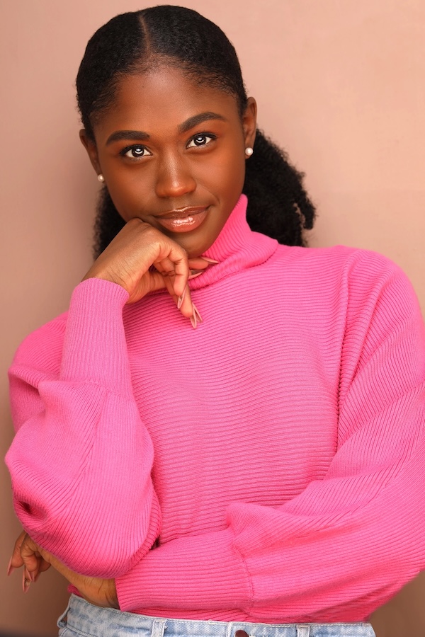 Woman in a bright pink turtleneck sweater smiles softly with her hand resting under her chin; neutral peach backdrop.
